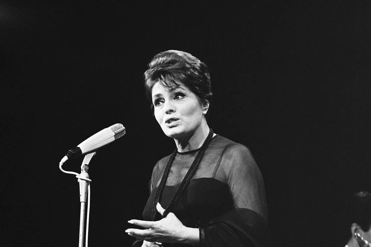 Black-and-white photograph of Amália Rodrigues singing on stage in a dark dress and shawl beside a microphone stand