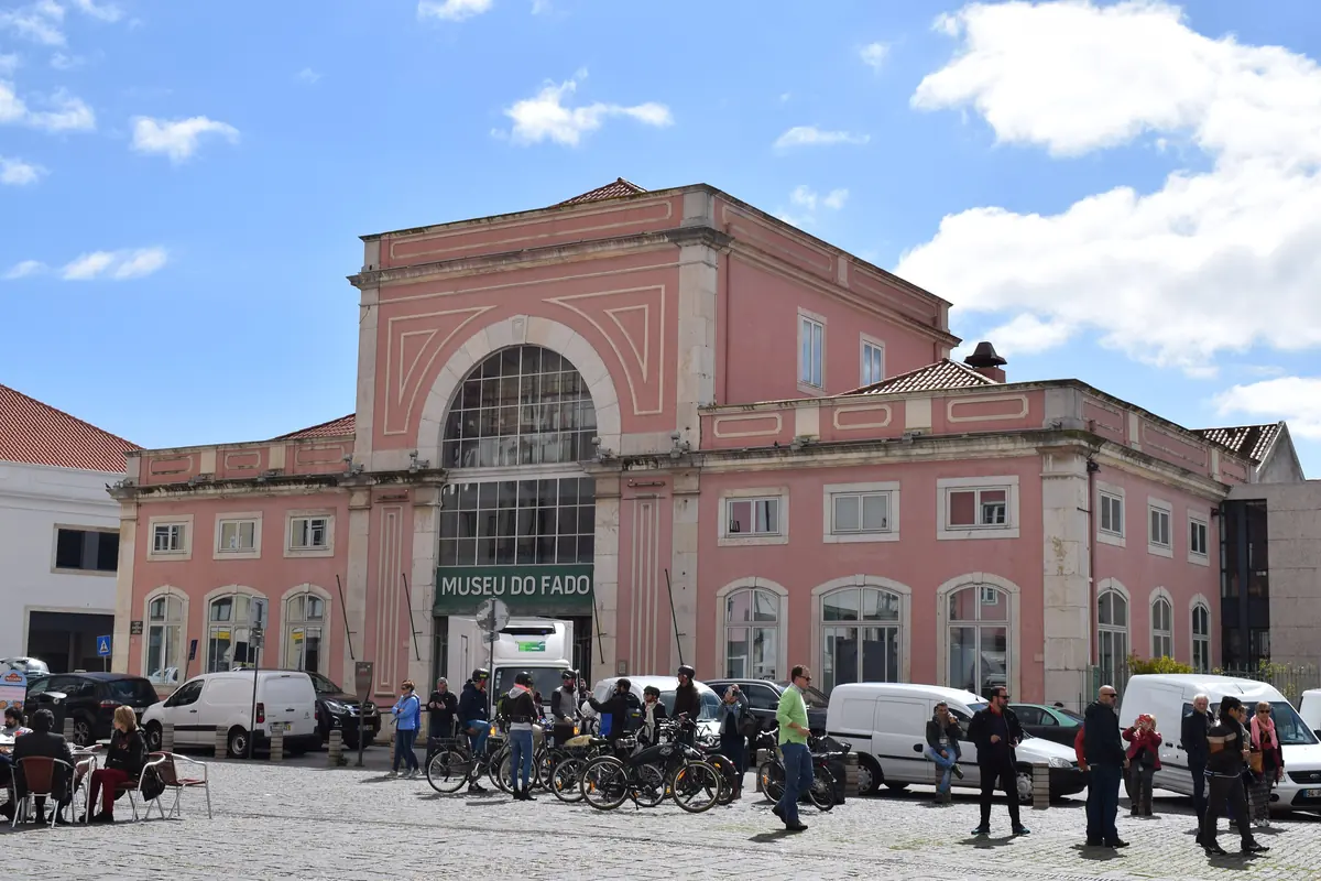 Pink neoclassical building of the Museu do Fado at the edge of a cobbled square, with people and parked vans in front and a blue sky behind